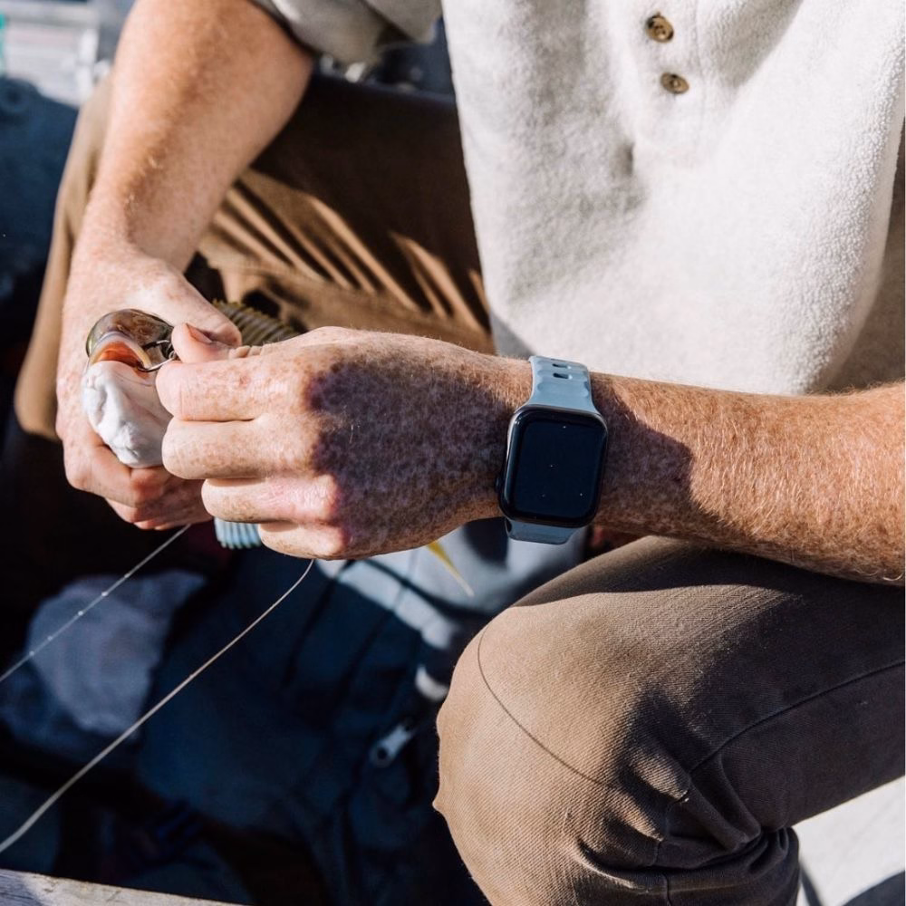 Ремешок Nomad Sport Band Marine Blue из прочного материала FKM на запястье, вид циферблата Apple Watch на фоне воды.